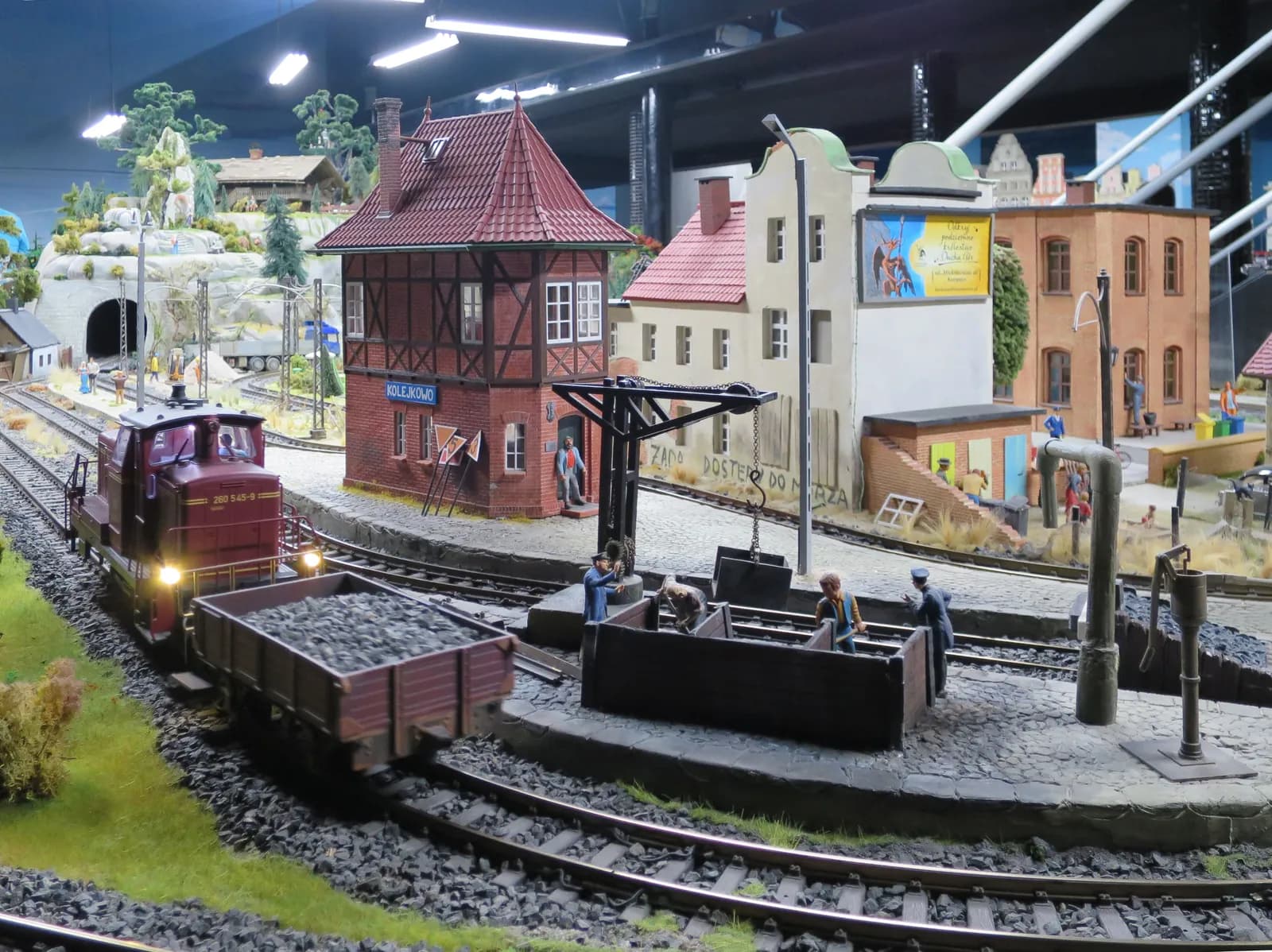 A miniature model train, loaded with coal, sits on tracks next to a miniature train station and buildings, with small figures around a crane.