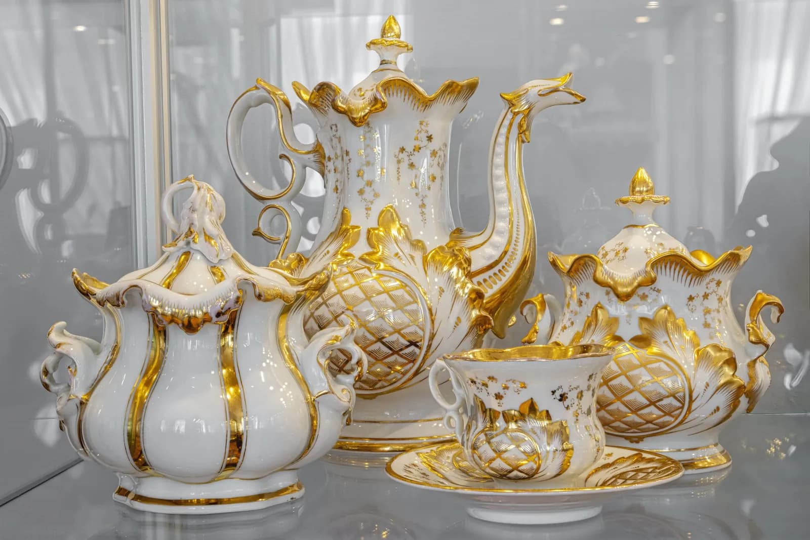A luxurious white and gold porcelain tea set is displayed, featuring a large teapot, sugar bowl, creamer, and teacup with a saucer, all intricately decorated with gold accents and a pineapple pattern.