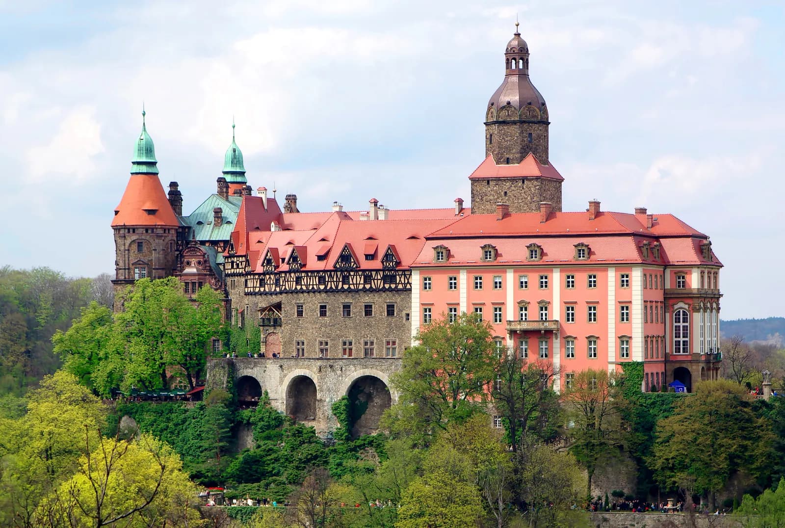 A grand, multi-colored castle with red and green roofs and a tall central tower, nestled among green trees under a partly cloudy sky.