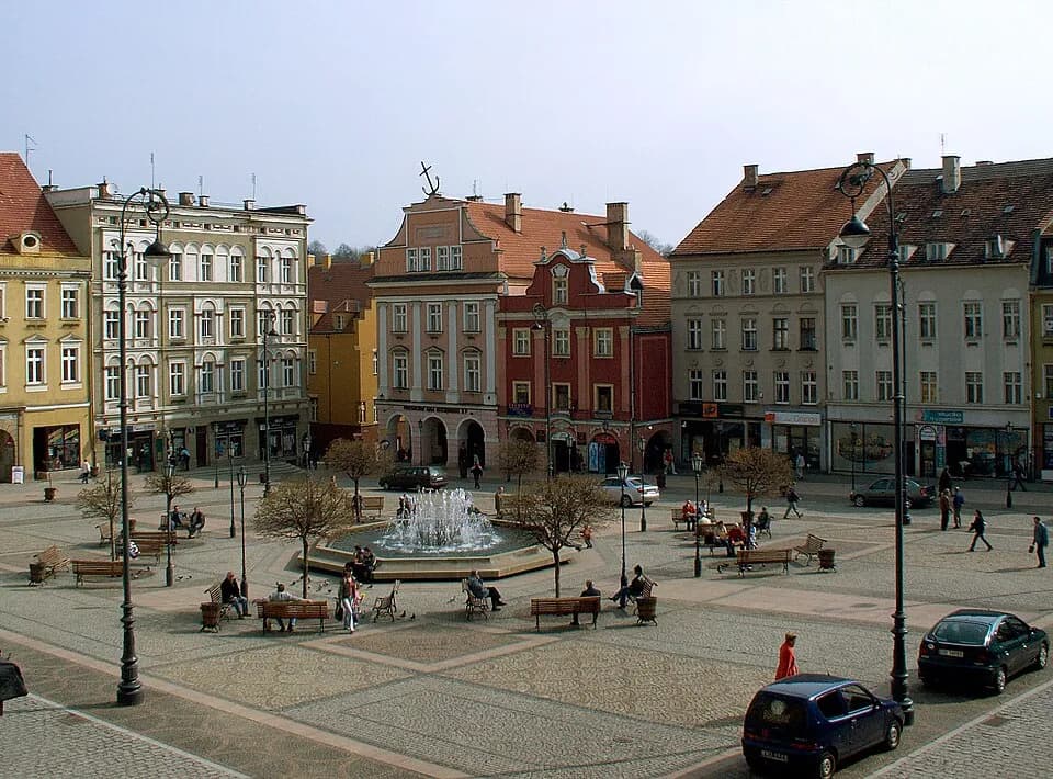 A large cobblestone town square features a central fountain with people sitting on benches around it, surrounded by multi-story historic buildings with tiled roofs under a clear sky.